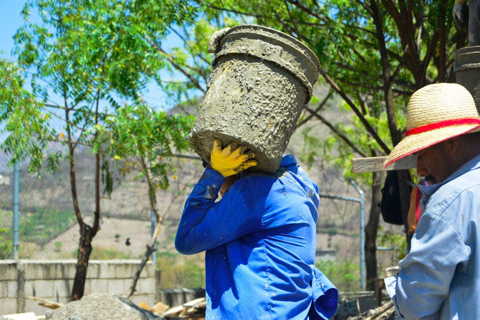 Construction worker carrying heavy concrete bucket under trees outdoors