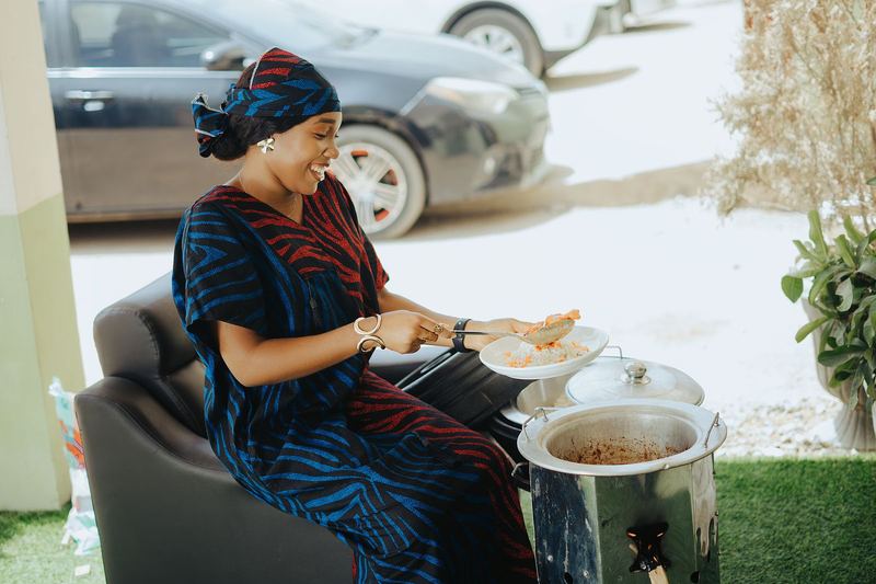 Smiling African woman cooking a traditional meal outdoors, serving food from a pot