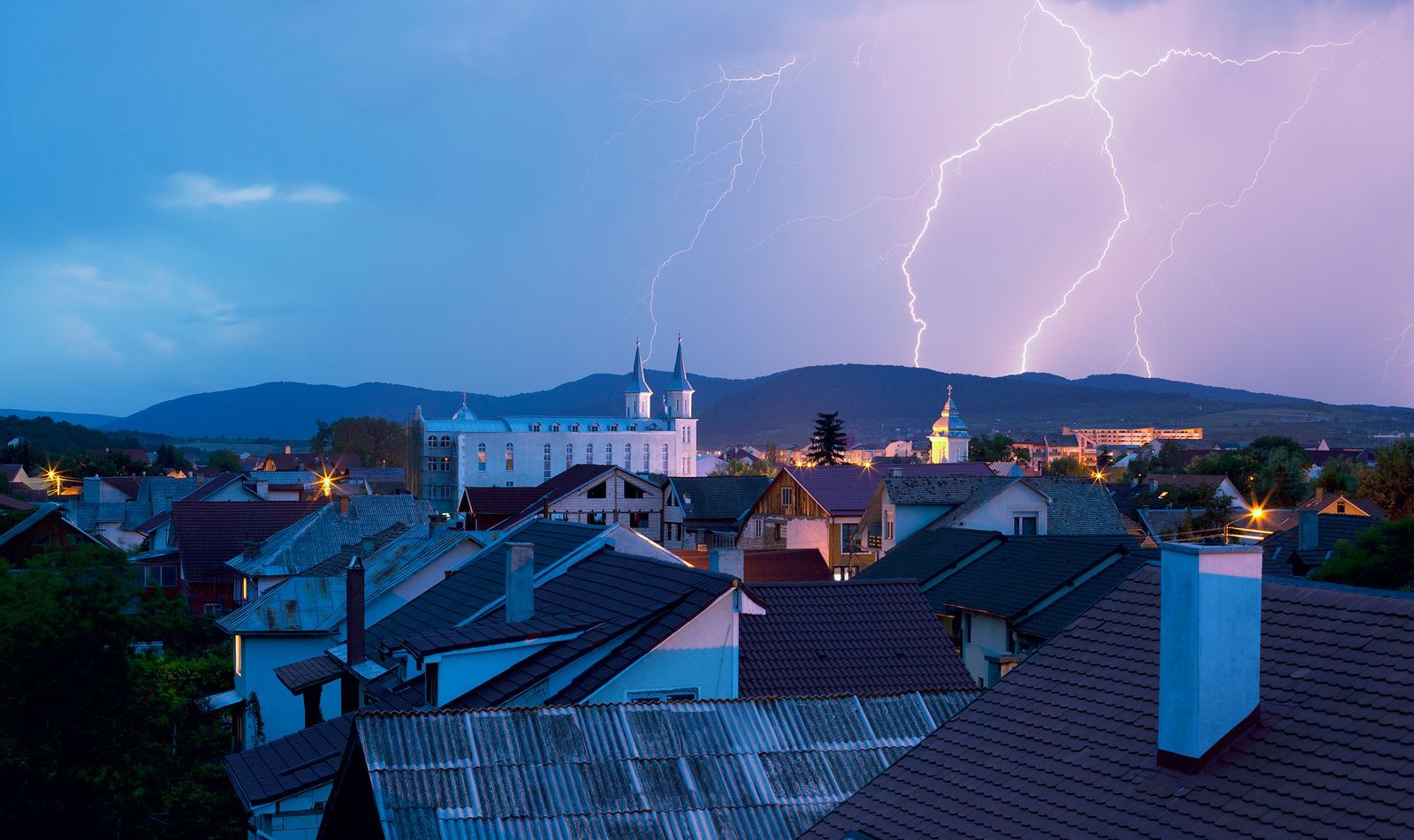 A powerful lightning storm illuminates the night sky over a town with rooftops and distant hills
