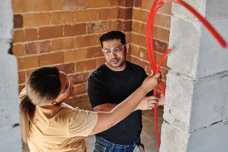 Two construction workers fitting red tubing on a brick wall at a construction site